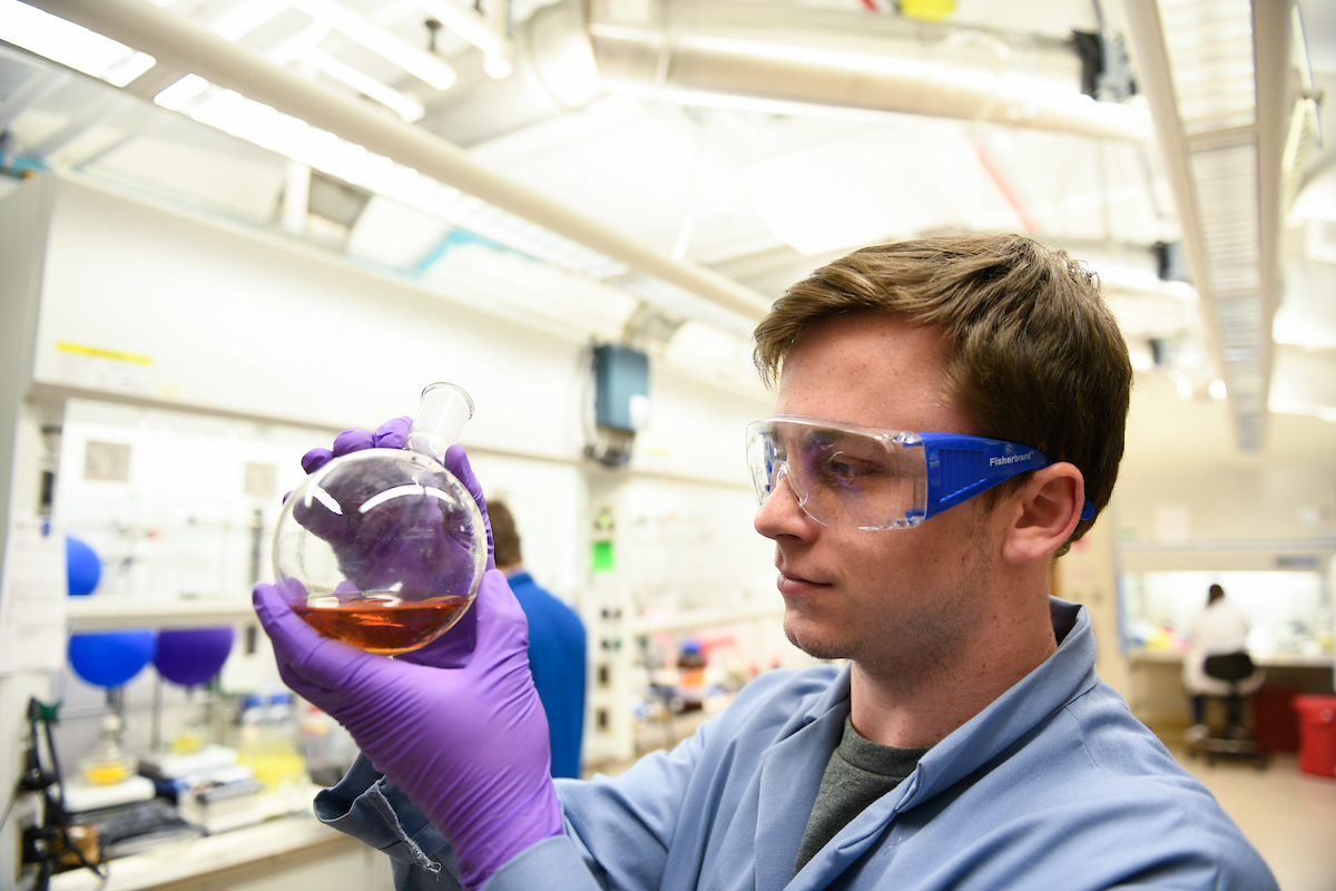 As an example of undergraduate research a chemistry student in a lab wearing safety gear is examining a flask with a dark-colored fluid inside.