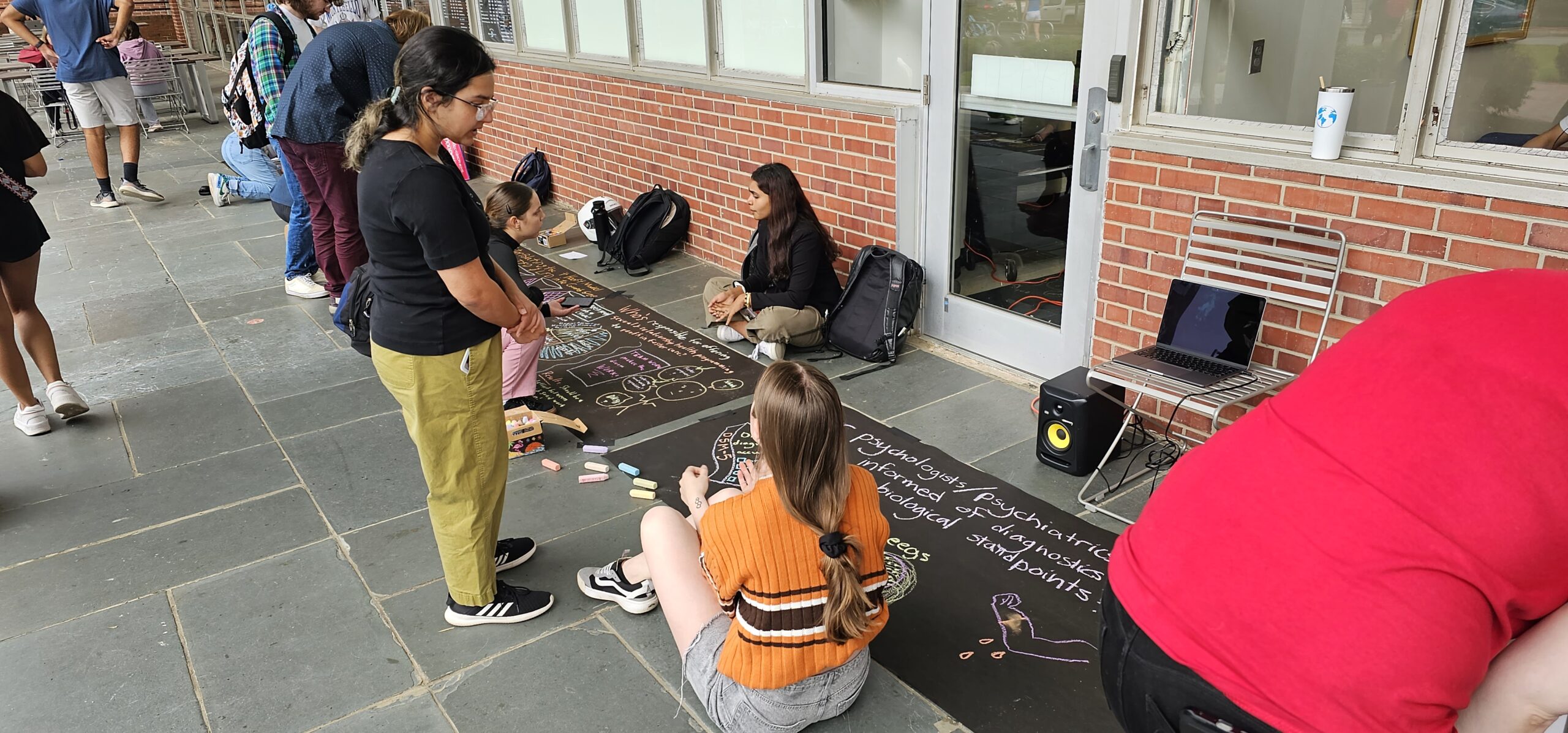 Students on the sidewalk creating drawings based on their research using chalk.  Visitors are standing nearby talking with student artists.